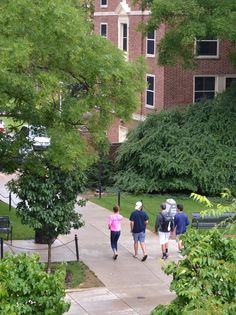 three people walking down a sidewalk in front of a brick building with trees and bushes