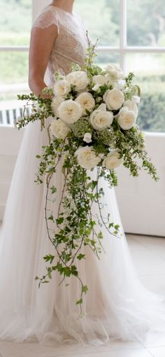 a woman in a wedding dress holding a bouquet of white roses and greenery on her shoulder