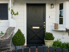 a black front door on a white house with two wicker chairs and potted plants