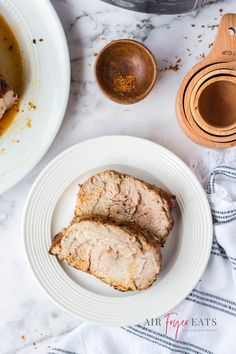 two pieces of meat on a white plate next to a bowl of soup and a wooden spoon