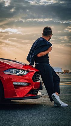 a man sitting on the hood of a red sports car in a parking lot at sunset