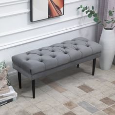 a gray bench sitting in front of a white vase and plant on the floor next to a window