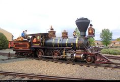 an old fashioned steam engine is on the train tracks with people standing on top and looking at it