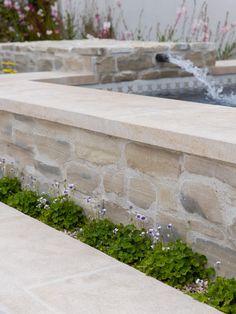 an outdoor fountain surrounded by plants and flowers on the side of a building with water coming out of it