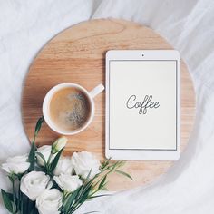 a cup of coffee next to a tablet on a wooden tray with white flowers and greenery