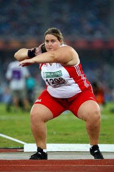 a woman in red shorts and white shirt throwing a ball on a track with her arms outstretched