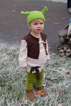 a little boy wearing a green and brown costume standing in the grass next to a stroller