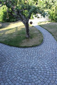 a brick pathway with a tree and swing in the background, surrounded by grass and trees