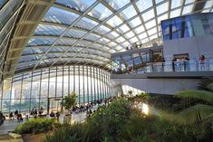 the inside of a building with lots of plants and people walking around it in front of large glass windows