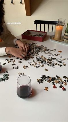 a woman sitting at a table working on a puzzle piece with two glasses next to it