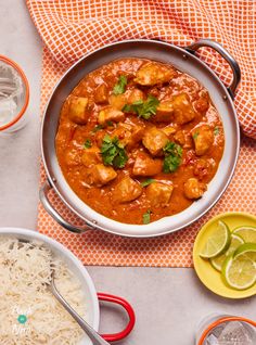 a pan filled with chicken curry next to rice and lime wedges on a table