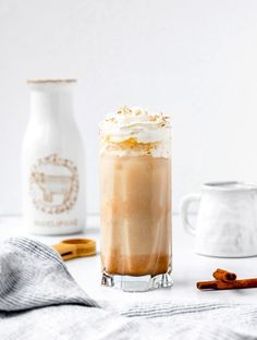 an iced drink with whipped cream and cinnamon sticks next to two milk jugs on a white table