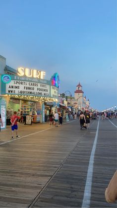 people are walking on the boardwalk near some shops and restaurants at dusk, in front of an oceanfront movie theater