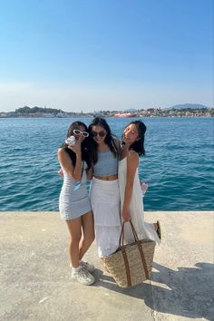 three young women standing next to each other near the water with one holding a purse