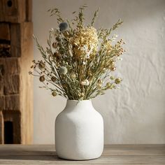 a white vase filled with lots of flowers on top of a wooden table next to a wall