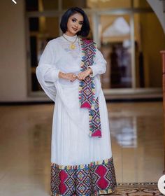 Ethiopian and Eritrean traditional dress,  elegant dress more vibrant and detailed embroidery. The patterns on the chest, cuffs, and hem are much more colorful, using a mix of red, yellow, green, black, and pink. The design on the neckline forms a prominent geometric motif, which is mirrored at the bottom of the dress and sleeves. The white fabric again serves as a canvas for these vivid details.  Any ceremony Habesha Dress Modern, Eritrean Dress, Beautiful Ethiopian, Ethiopian Clothing, Habesha Dress, Ethiopian Traditional Dress, Cultural Fashion, Ethiopian Dress, Habesha Kemis