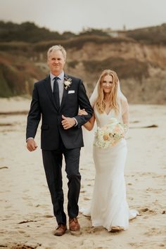 a bride and groom walking on the beach holding each other's hand as they walk towards the camera