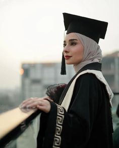 a woman wearing a black graduation gown and holding a wooden board in her hand while standing on top of a roof