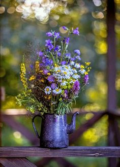 a watering can filled with wild flowers on top of a wooden table in front of trees