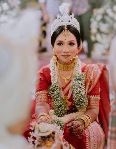 a woman in a red and gold wedding outfit sitting down with flowers around her neck