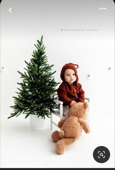 a baby sitting on a chair next to a small christmas tree with a teddy bear