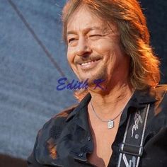 an older man with long hair smiling and wearing a black shirt in front of a blue background