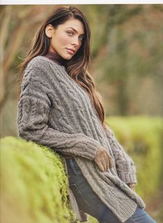 a woman leaning against a mossy wall wearing a gray sweater and jeans with her hands on her hips