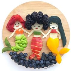 three little mermaids made out of fruit on a wooden plate with blueberries and raspberries