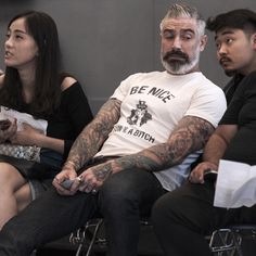 three people sitting next to each other with tattoos on their arms and legs, one man is looking at the camera