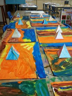 several colorful paintings are on the table with paper pyramids in them and children's books nearby