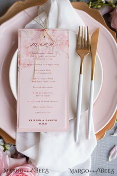 a pink and gold place setting with menu cards, silverware and flowers on the table