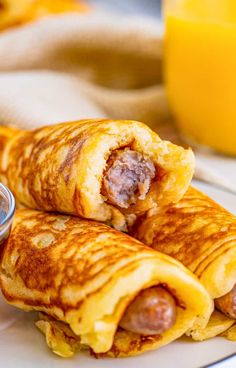 two sausage wrapped in crepes on a white plate next to a glass of orange juice