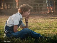 a young man sitting in the grass with his hand on his chin looking at something