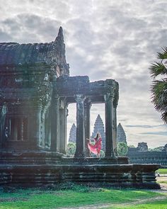 a woman in a red dress standing on the ruins of an ancient temple with palm trees