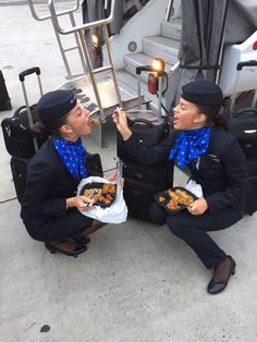 two women sitting on the ground eating food from their hands and holding onto luggage bags