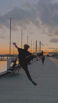 a person jumping in the air on a sidewalk near water and boats at sunset or dawn