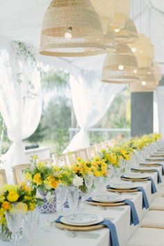 a long table with yellow and white flowers in vases on top of it, surrounded by hanging lights