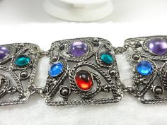 "Panel link bracelet with colorful gem acrylic stones in fancy open work metal. With four softly squared panels finished in silver tone metal, this statement link bracelet is simply beautiful. Each panel has a mix of purple, blue, green and red acrylic cabochons, some are domed, some are faceted. A twisted rope design wraps each stone as well as the outer the outer edges of each panel. Two \"0\" rings attach to each and connect each link. Measures 7 inches long and 1 3/8 inches wide. The bracelet closes with a sturdy wide fold over clasp. This piece is in excellent excellent condition and will be shipped in a new white gift box and ready for a perfect gift. RedD7 Hello and thank you for visiting CartersJewels. If you have any questions about an item, please contact me and I'll be happy to Retro Multicolor Metal Bracelets, Funky Jewelry, Rope Design, White Gift Boxes, Green And Red, Simply Beautiful, Chain Link Bracelet, Link Bracelets, Chain Link