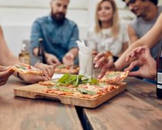 several people sitting around a wooden table eating pizza and drinking beer, with one person reaching for the slice