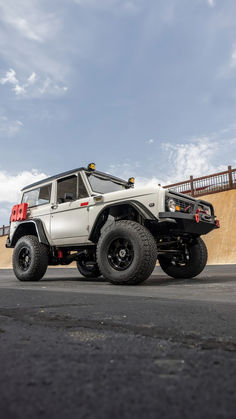 A white first-generation Ford Bronco with an LS engine swap, viewed from a low angle against a bright sky with scattered clouds. The vehicle features yellow roof lights, a black grille, winch, and off-road tires, conveying a bold and adventurous spirit Old Ford Trucks, Ls Swap