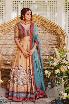 a woman standing in front of a wicker chair wearing a beige and blue lehenga