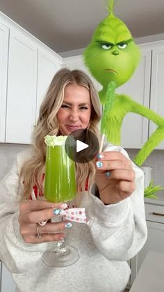 a woman holding up a green drink in front of an image of the grin face