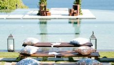 an empty bench with pillows on it in front of a pool and some water features