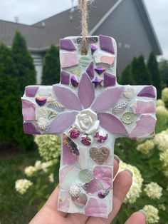 a hand holding a cross made out of stained glass and buttons, with a house in the background