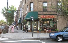 people are walking down the sidewalk in front of shops on a city street with parked cars