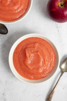 two bowls filled with red sauce next to an apple and spoon on a white surface