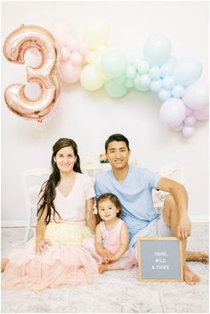 a man, woman and child sitting on a bed in front of balloons with the number three