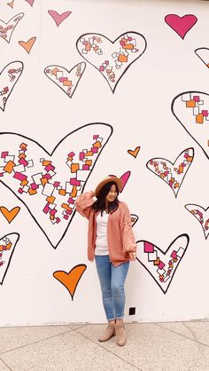 a woman standing in front of a wall with hearts painted on it and wearing a hat