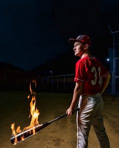 a young man holding a baseball bat on top of a field at night with flames in the air