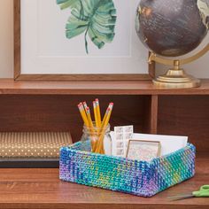 a wooden desk topped with a blue basket filled with pens and pencils next to a globe